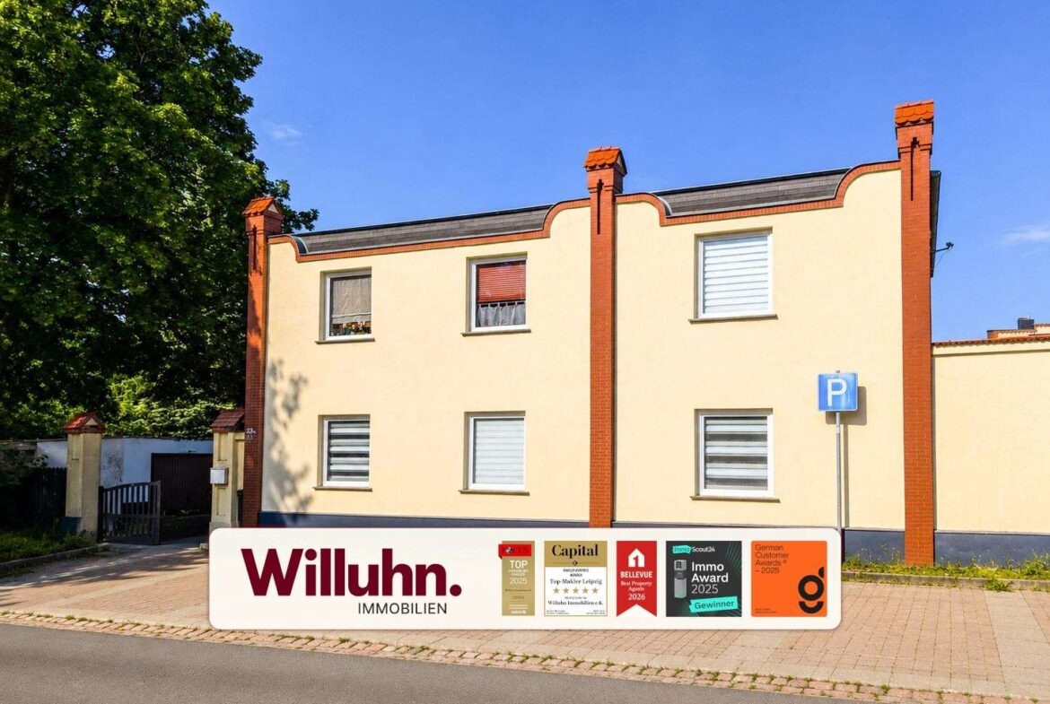 Two-story beige building with red brick trim and a clear blue sky; Willuhn Immobilien sign and award plaques in front.