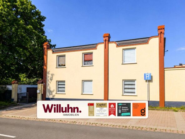 Two-story beige building with red brick trim and a clear blue sky; Willuhn Immobilien sign and award plaques in front.