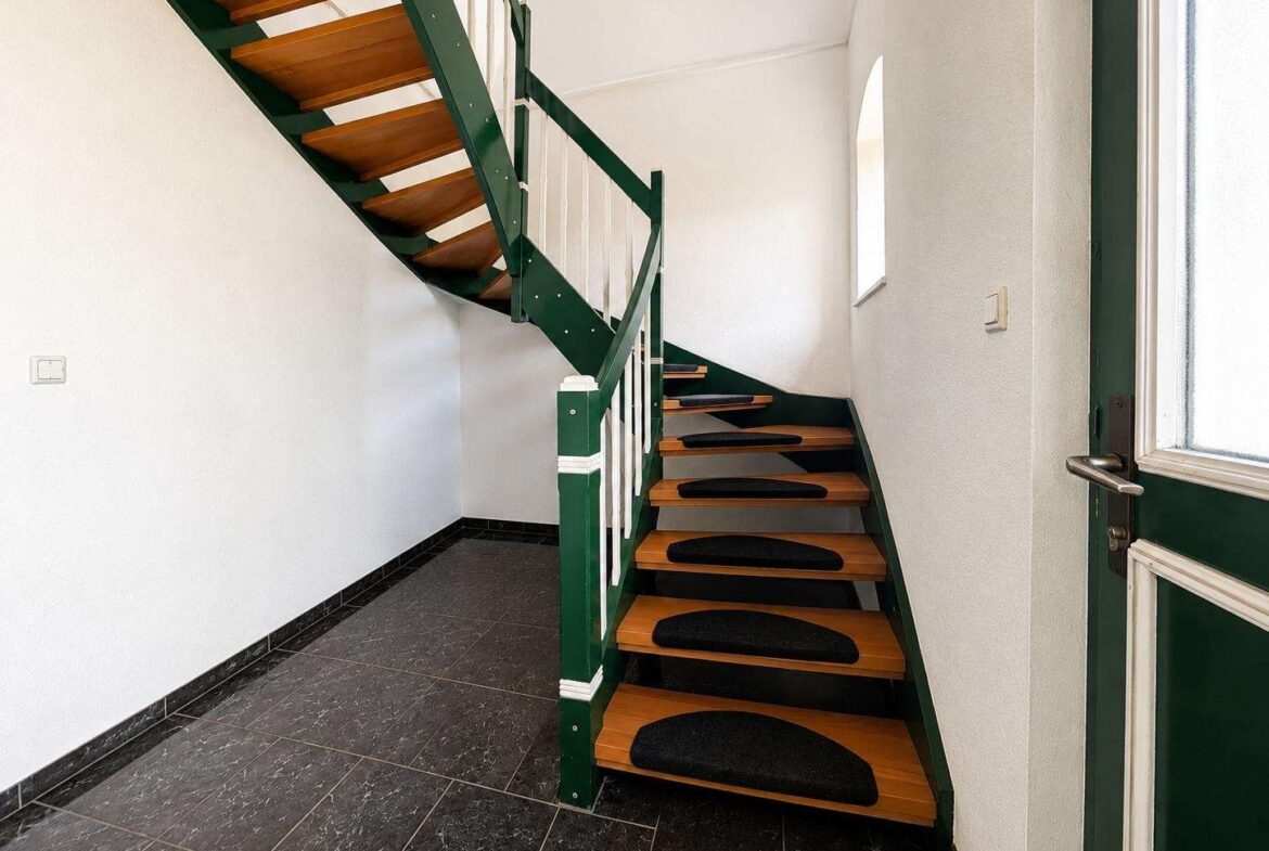 Interior entry hall with a two-flight wooden staircase, green metal railing, and white walls; black non-slip mats on the steps.