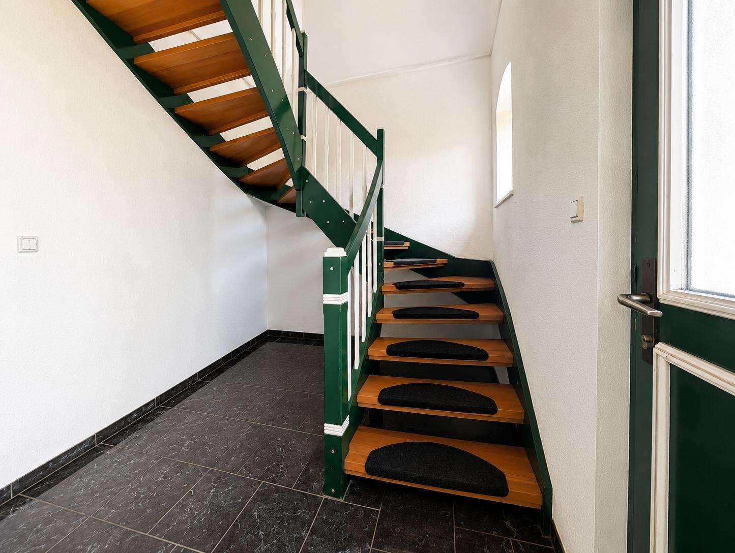 Interior entry hall with a two-flight wooden staircase, green metal railing, and white walls; black non-slip mats on the steps.