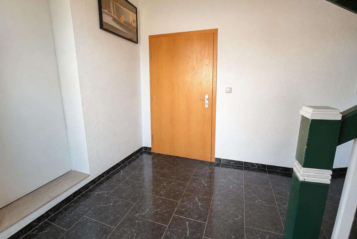 Interior hallway with a light wood door on white textured walls, dark marble floor tiles, a framed artwork on the left, and a green stair railing on the right.