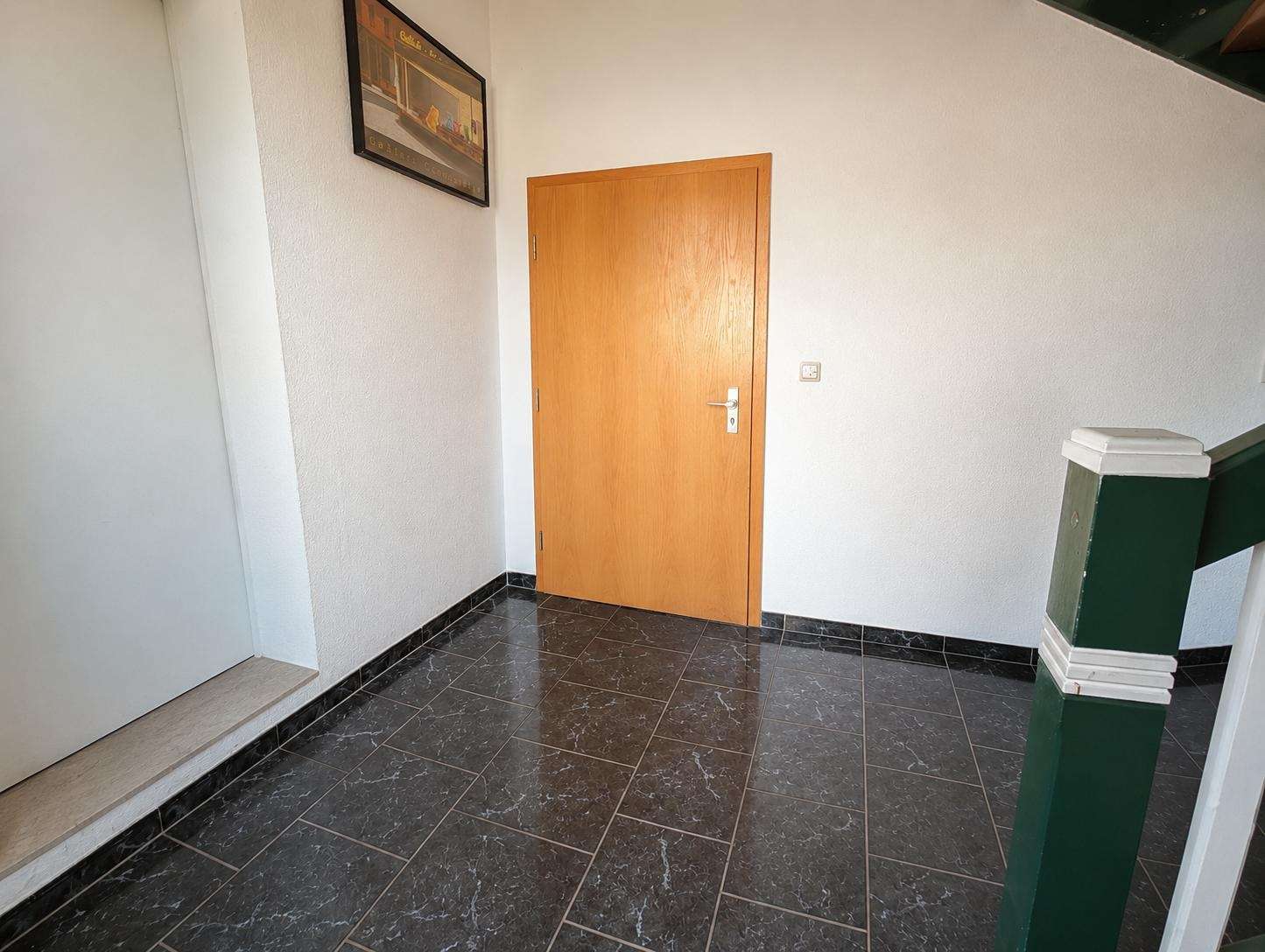 Interior hallway with a light wood door on white textured walls, dark marble floor tiles, a framed artwork on the left, and a green stair railing on the right.