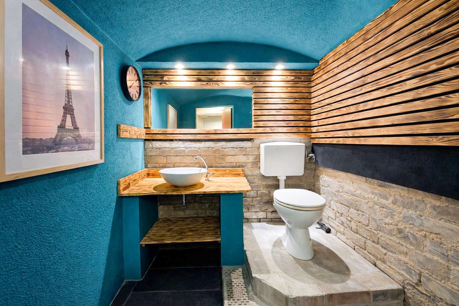 Teal-walled bathroom with a wooden vanity, vessel sink, and brick accents; toilet on a raised concrete platform against a brick wall and slatted wood panels above.