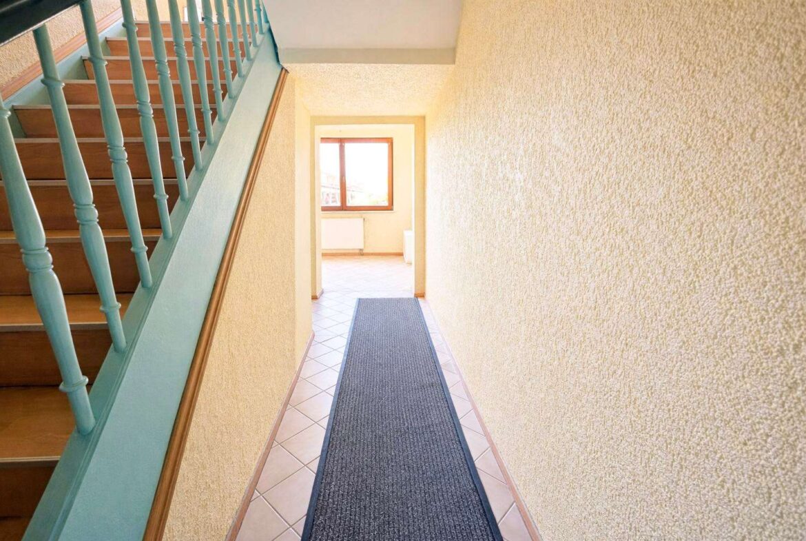 Narrow hallway with a blue runner rug, pale yellow textured walls, and a wooden staircase with turquoise balusters on the left; bright window at the far end.