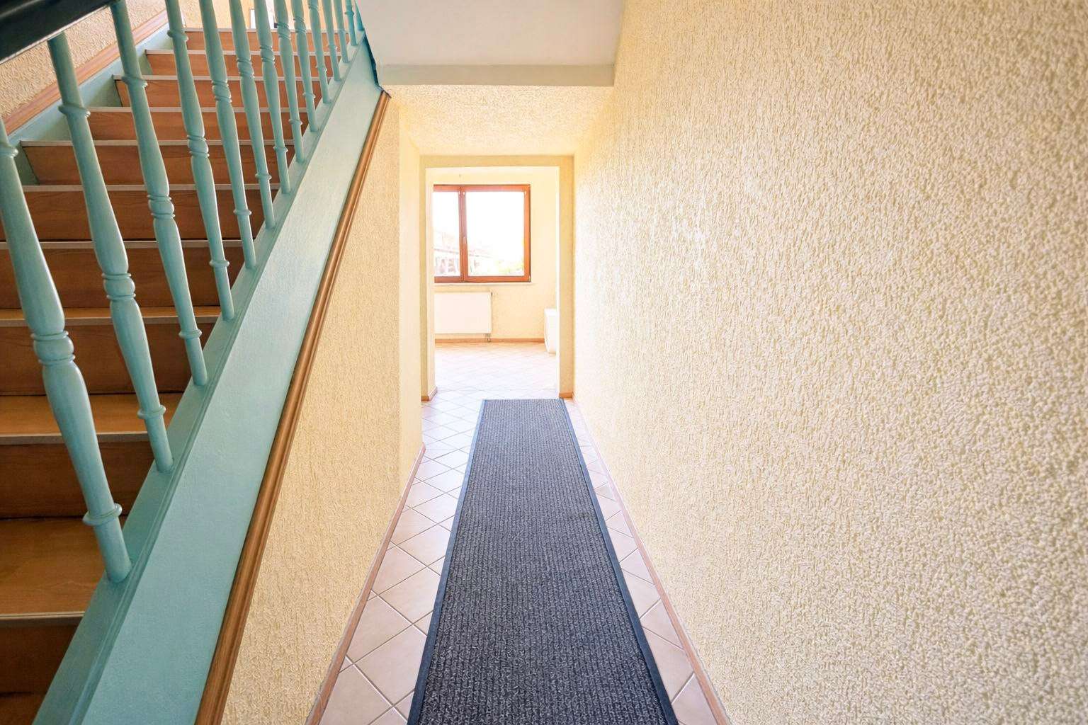 Narrow hallway with a blue runner rug, pale yellow textured walls, and a wooden staircase with turquoise balusters on the left; bright window at the far end.