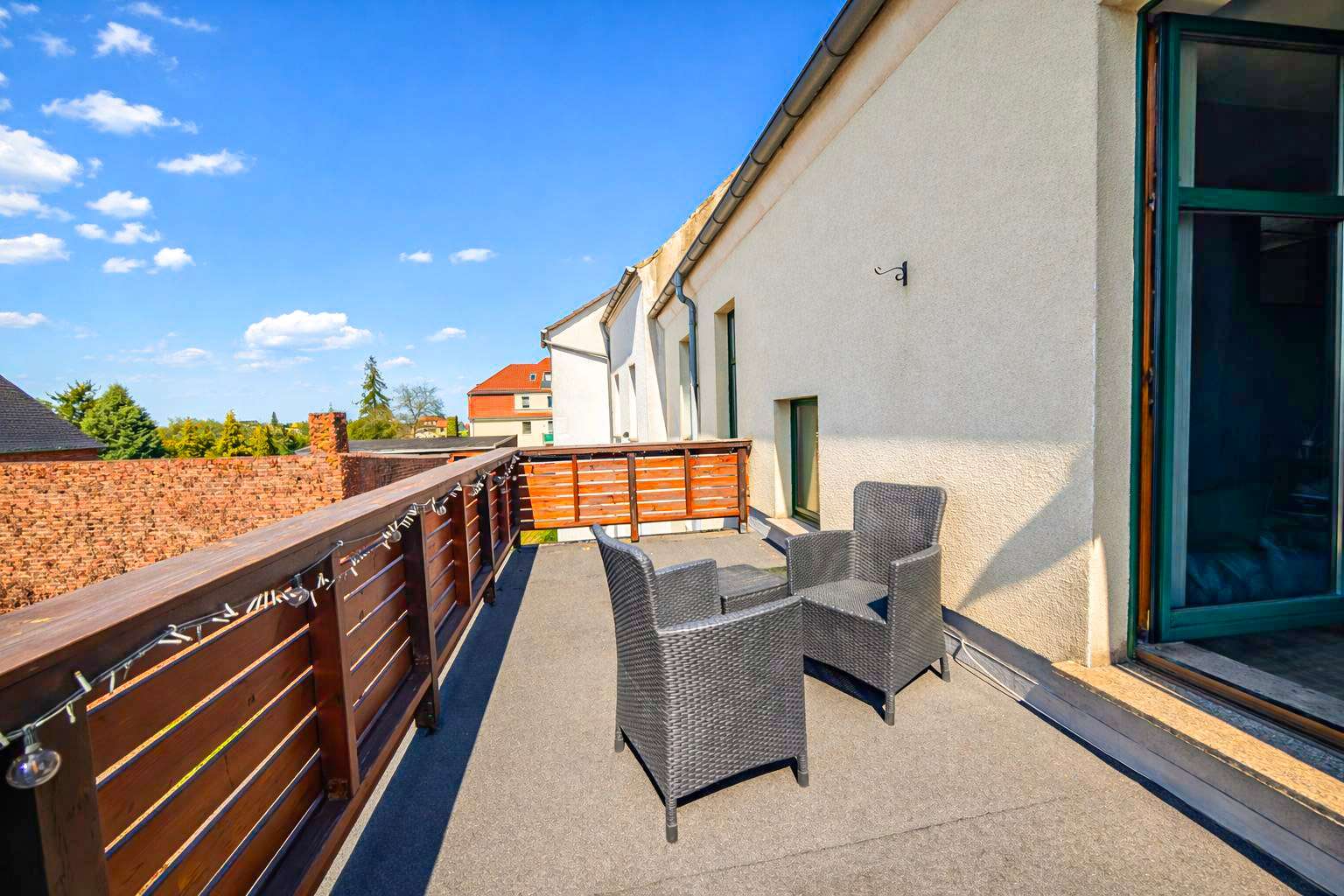 Rooftop terrace with two dark wicker chairs and a small table, wooden railing, bright blue sky, string lights along the railing.