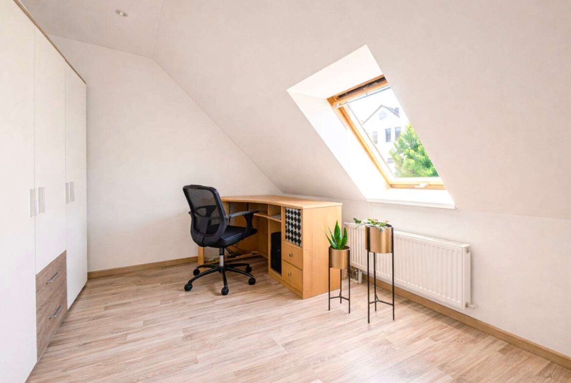 Home office in an attic with a sloped wall, wooden desk, black mesh chair, white wardrobe, and a skylight window bringing in natural light.