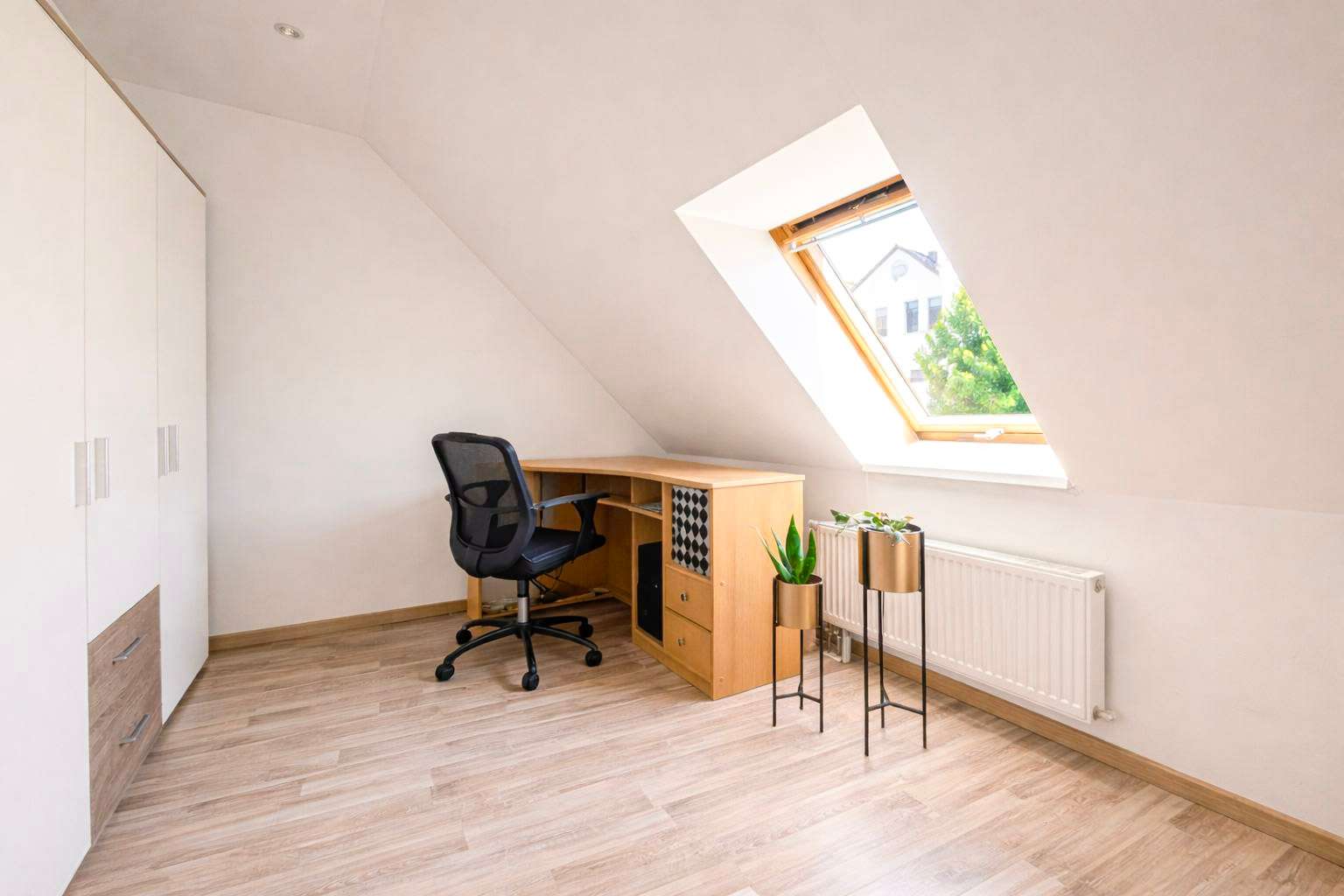 Home office in an attic with a sloped wall, wooden desk, black mesh chair, white wardrobe, and a skylight window bringing in natural light.