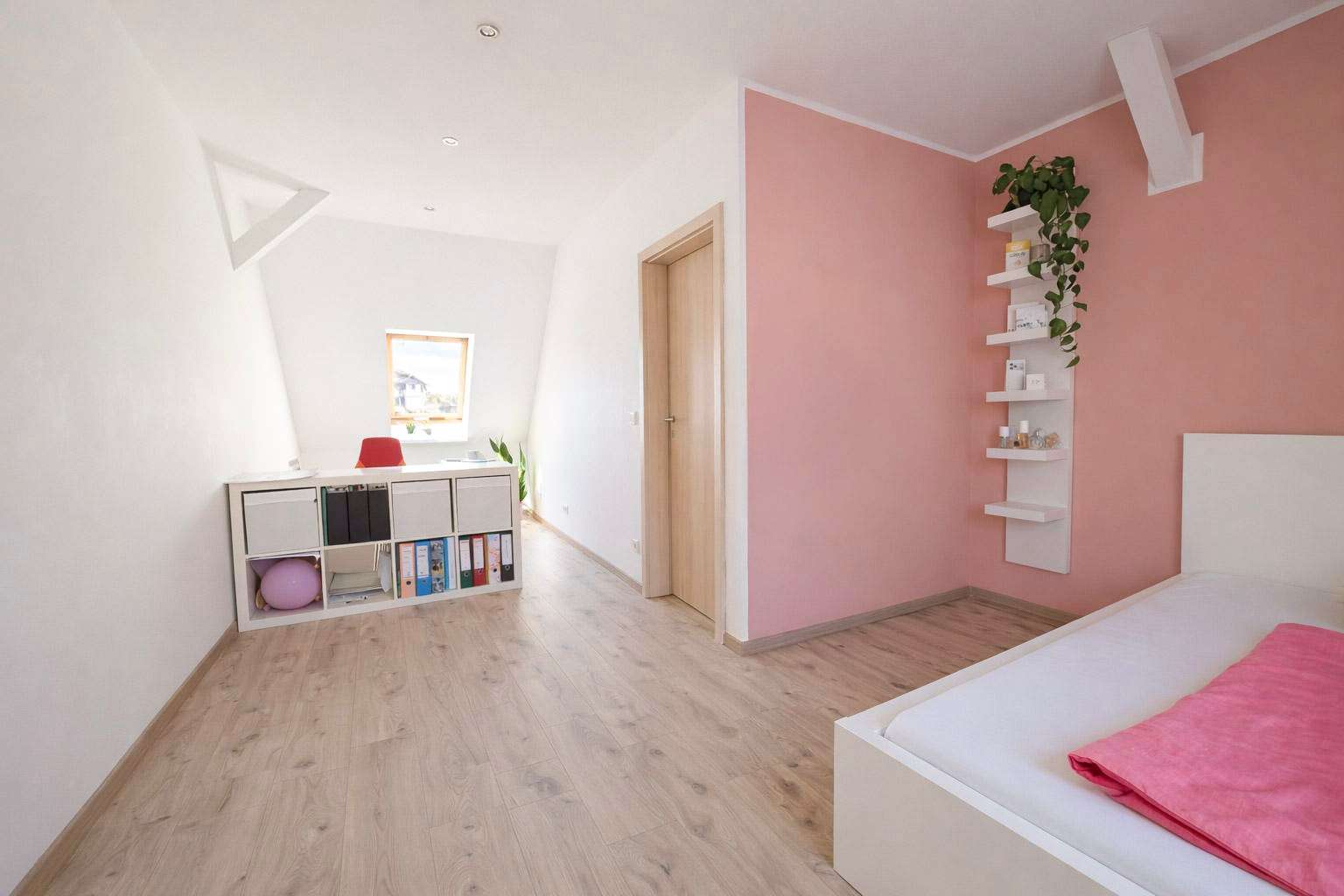 Bedroom with pink accent wall, white bed and floating shelves, light wood floor, and a desk area near a window. A red chair sits at the desk.