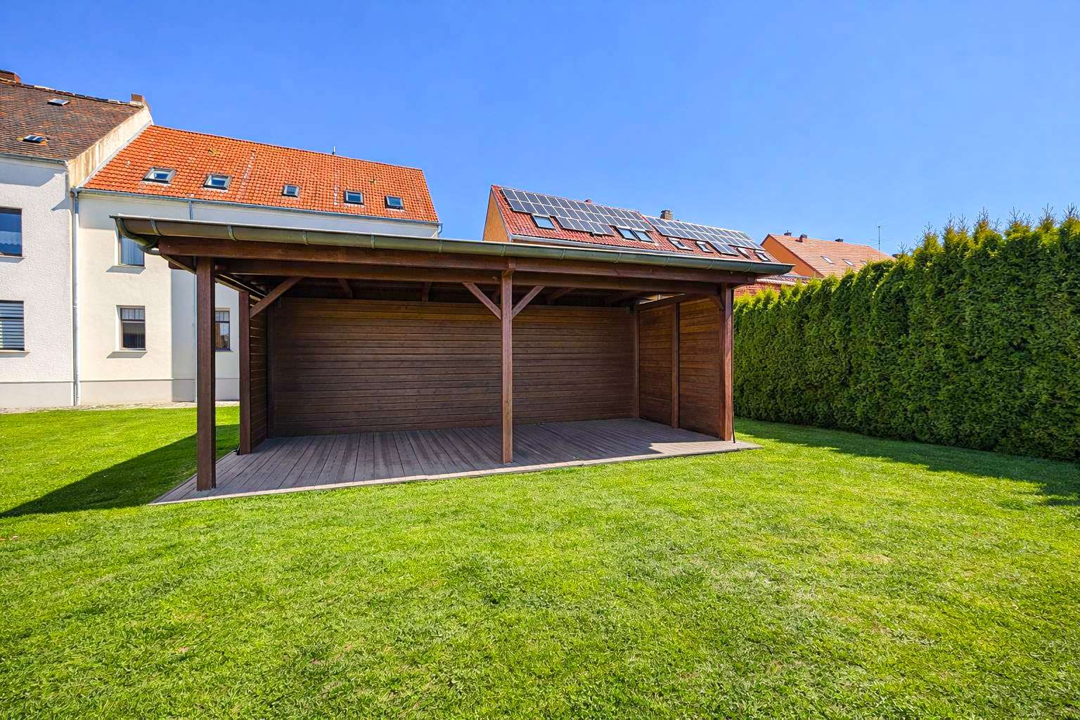 Wooden carport with a green lawn, attached to a white house, under a clear blue sky; solar panels on the neighboring roof.