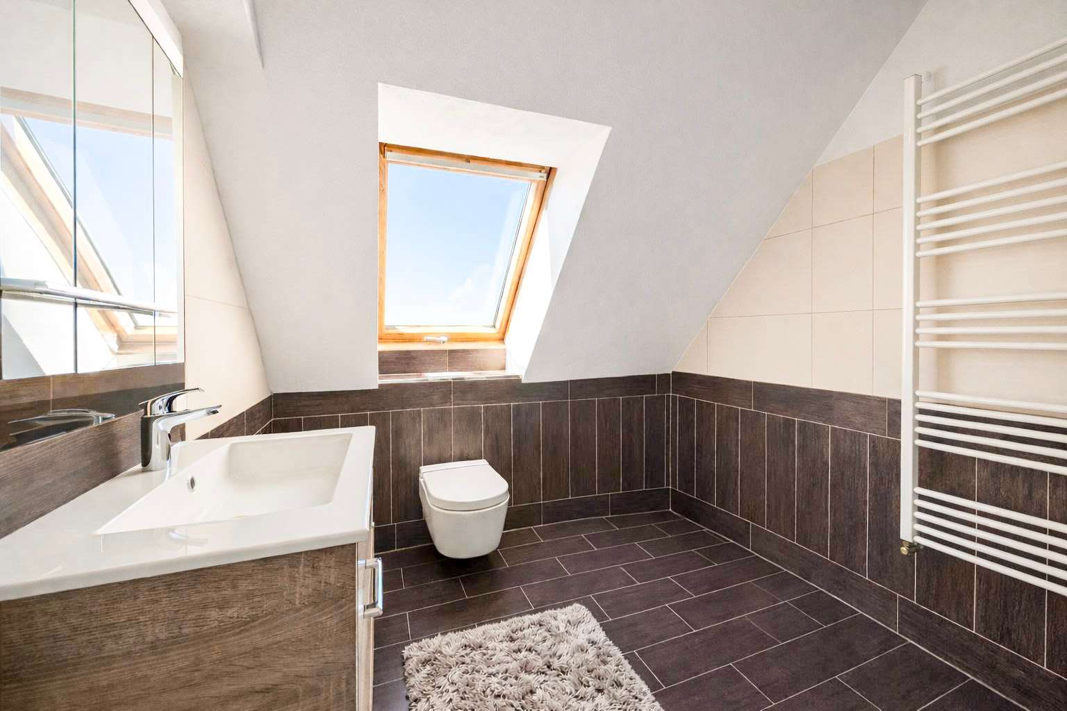 Bathroom with a wall-hung toilet, a white sink, and a skylight window letting in natural light; dark brown floor and wall tiling with a fluffy rug on the floor.