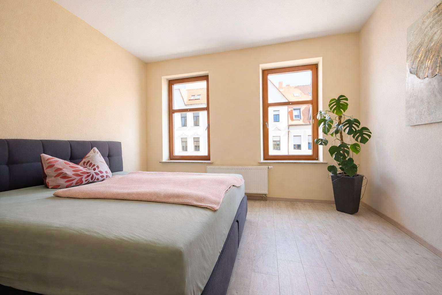 Bright bedroom with a low, upholstered bed and pink throw pillows by two wooden-framed windows; pale walls and light floor.