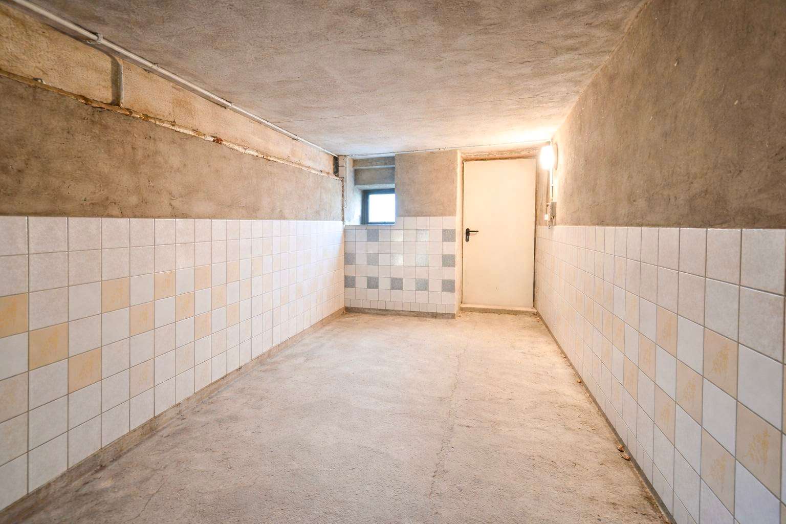 Long concrete basement corridor with tiled lower walls and a white door at the far end, small window beside it