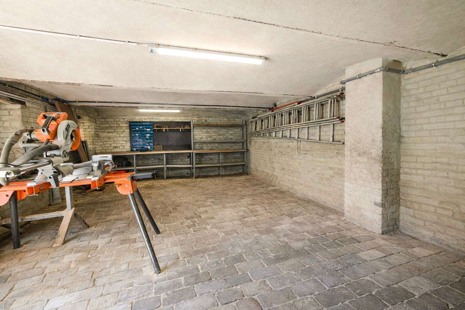 Basement workshop with a miter saw on a stand, workbench, and metal shelves against brick walls.