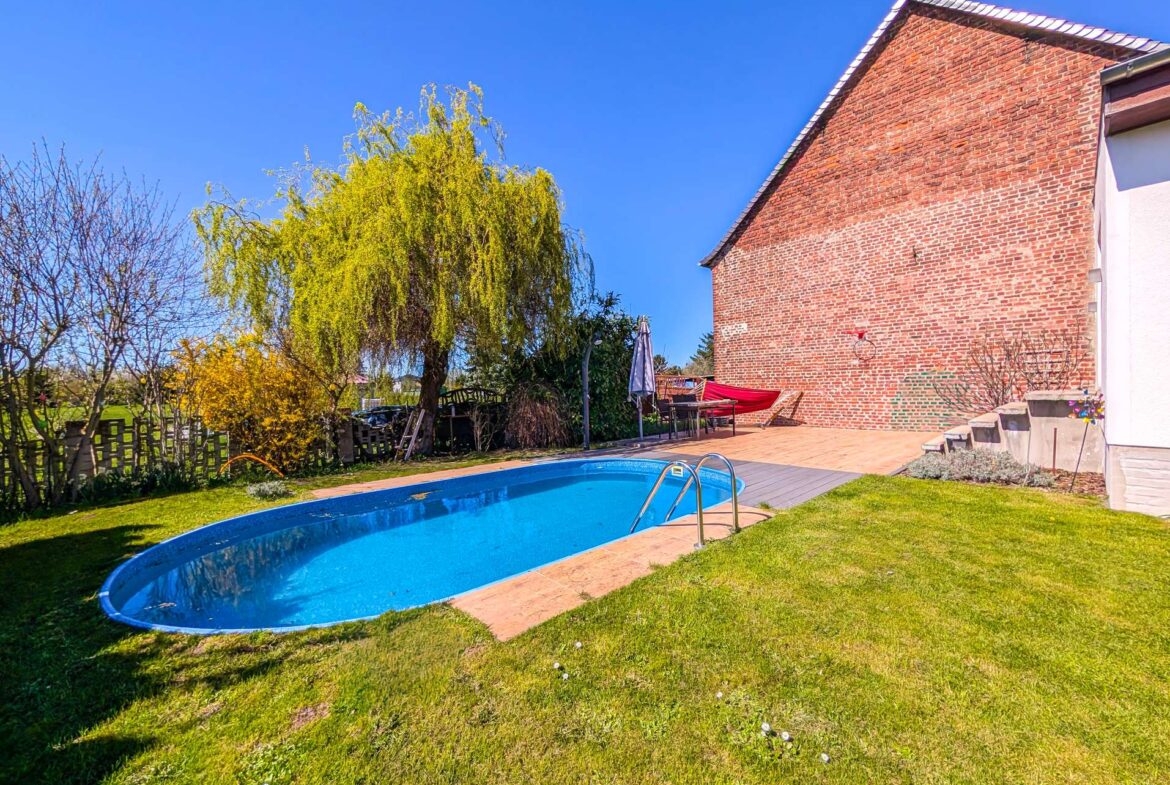 Backyard with a blue in-ground pool, tiled decking, and a brick house with a sloped roof under a clear blue sky.