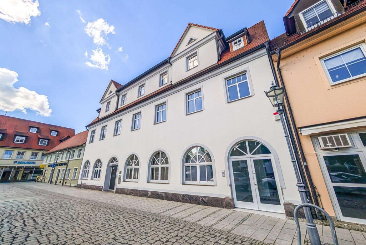 White three-story building with arched ground-floor windows on a cobblestone street under a blue sky, with neighboring peach building to the right.