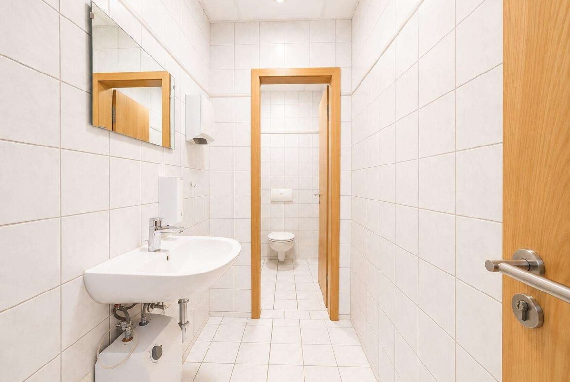 Bright white tiled bathroom with a wall-mounted sink, mirror, and toilet visible through an open doorway at the back of the room.