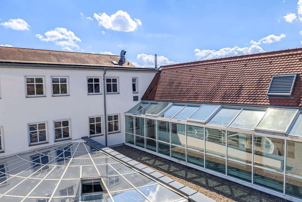 Courtyard scene showing a white building with multiple windows and a glass-roofed atrium covering a gravel area under a blue sky with clouds.