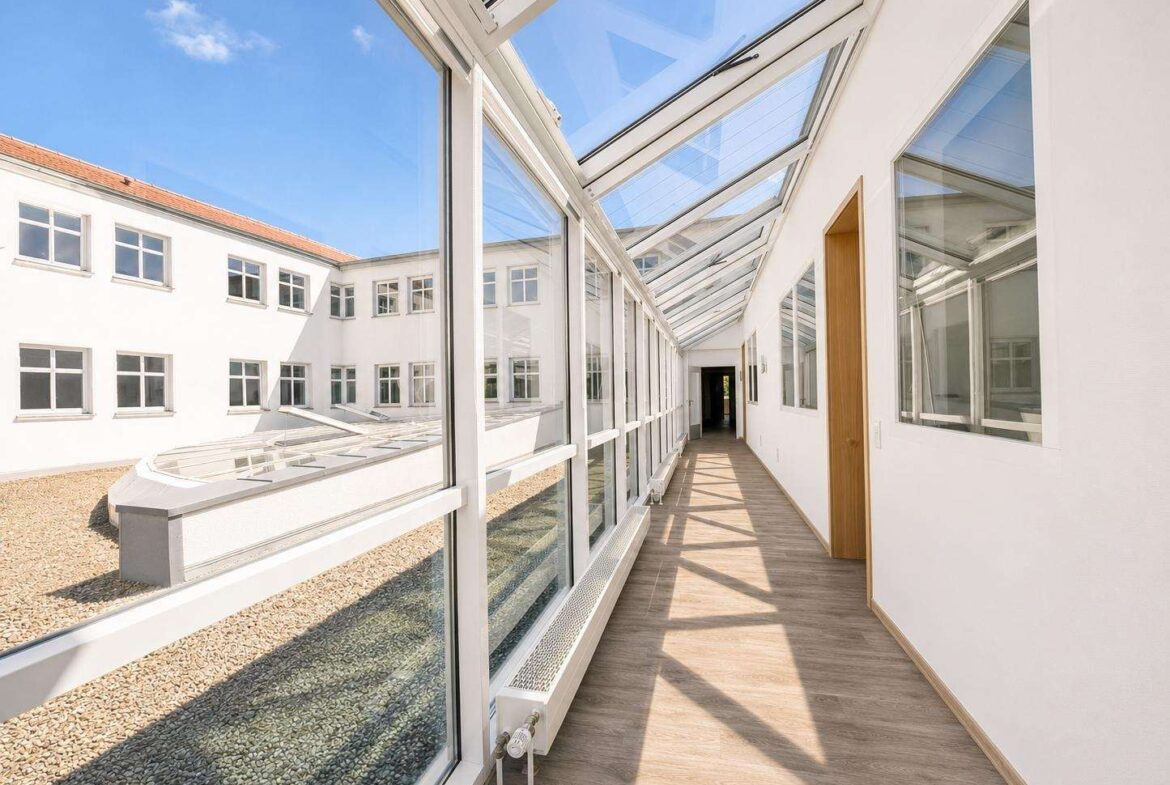Bright glass-roofed corridor inside a white building, with a wooden door on the right and sunlight casting shadows on the wooden floor.