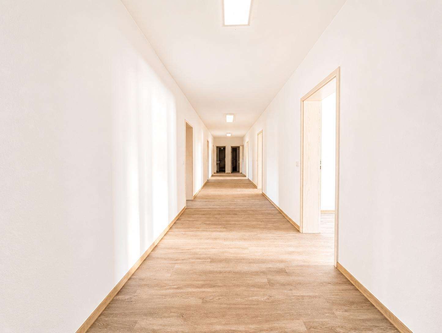 Bright white hallway with light wood flooring and doors along both walls, extending toward a distant doorway.