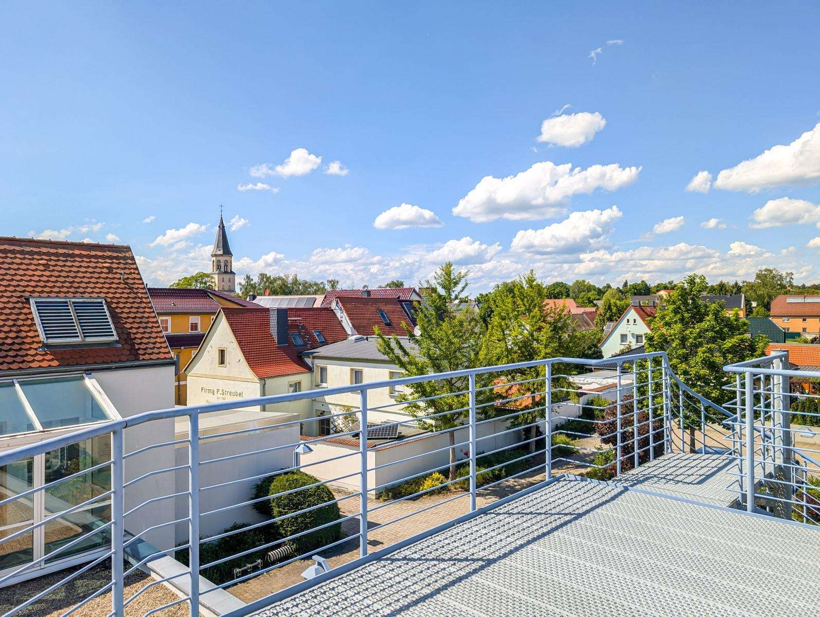 Rooftop terrace with metal railing overlooking a small European town with red-tiled roofs and a church steeple on the horizon under a blue sky.