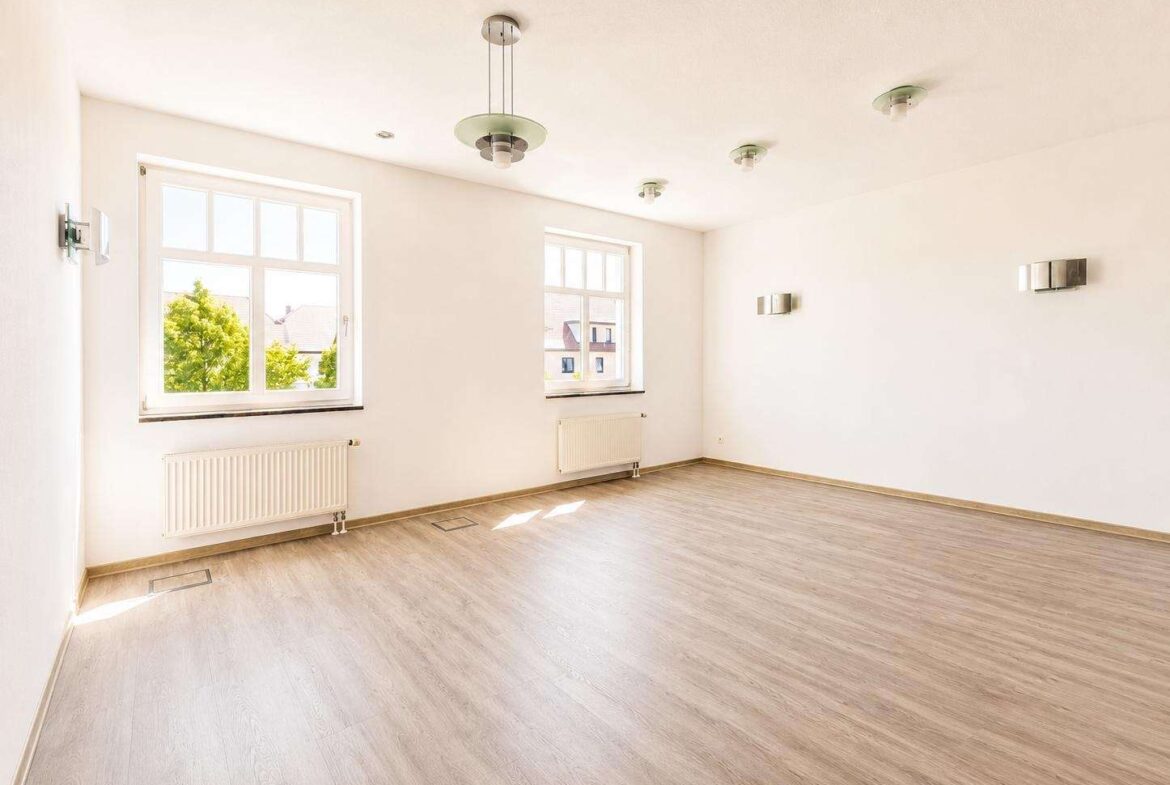 Bright, empty living room with two large windows, white walls, and light wood flooring. Ceiling lights and radiators visible.