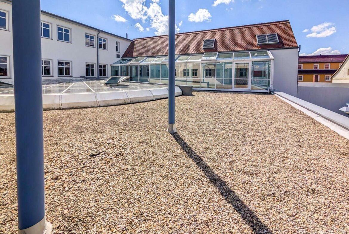 Rooftop gravel terrace with glass skylights and a white building under a bright blue sky.