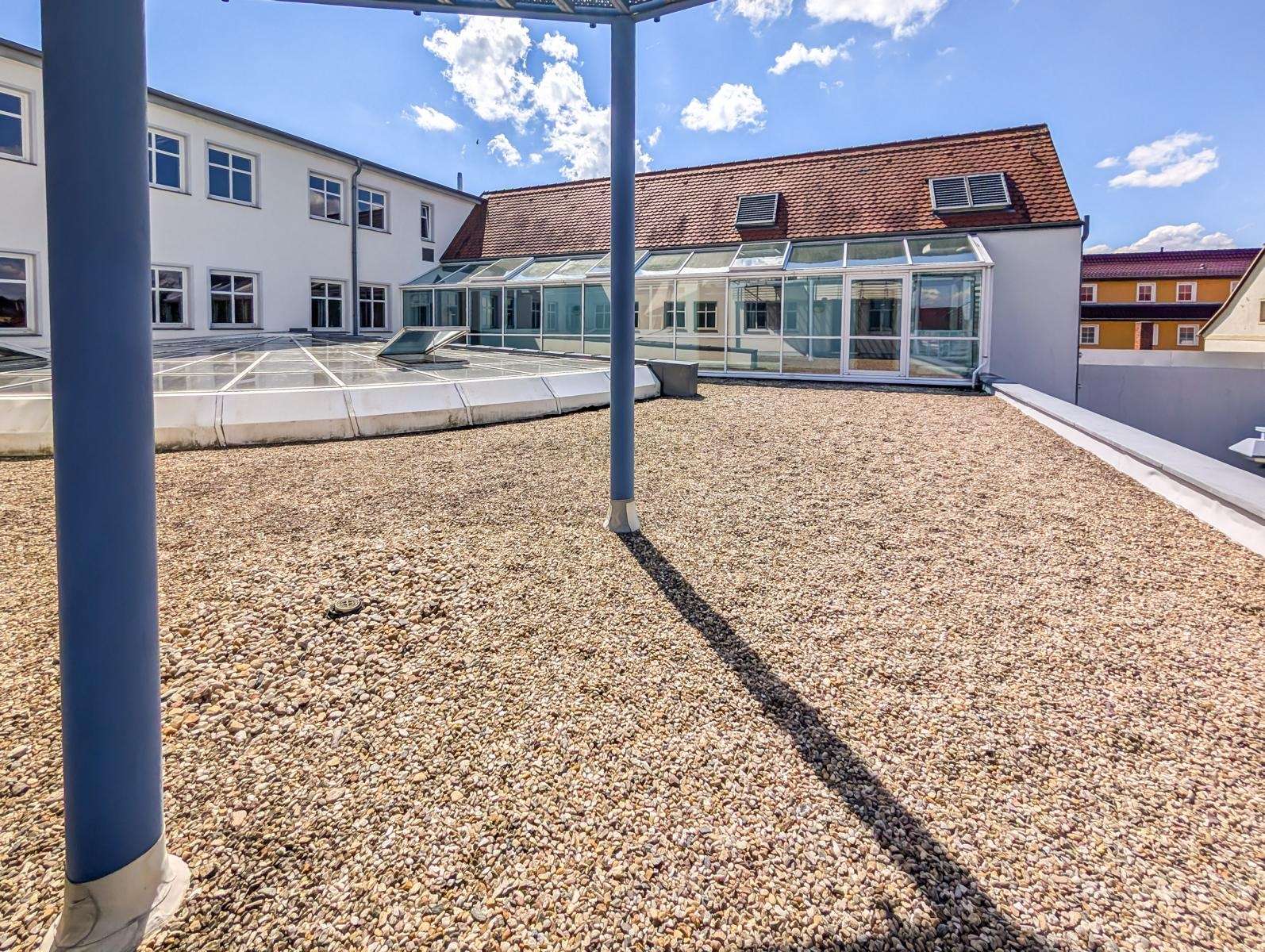 Rooftop gravel terrace with glass skylights and a white building under a bright blue sky.
