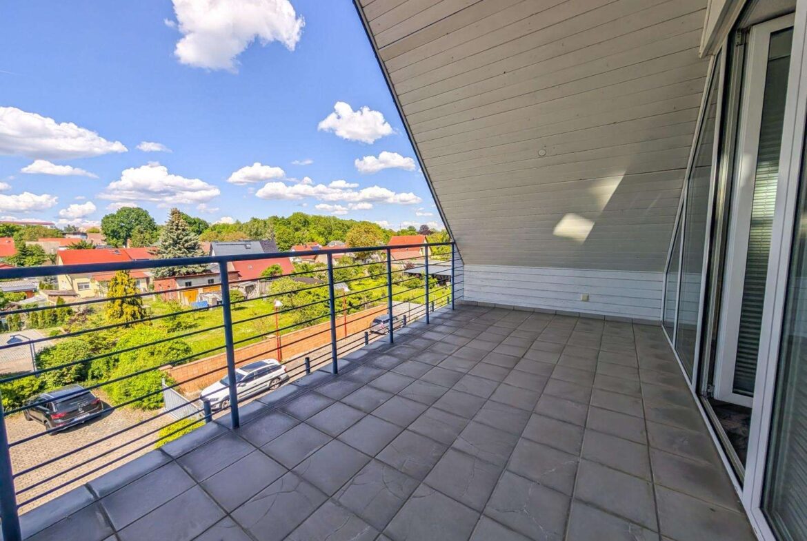 Empty balcony with gray tiled floor and blue metal railing overlooking a suburban neighborhood under a sunny blue sky with fluffy clouds.