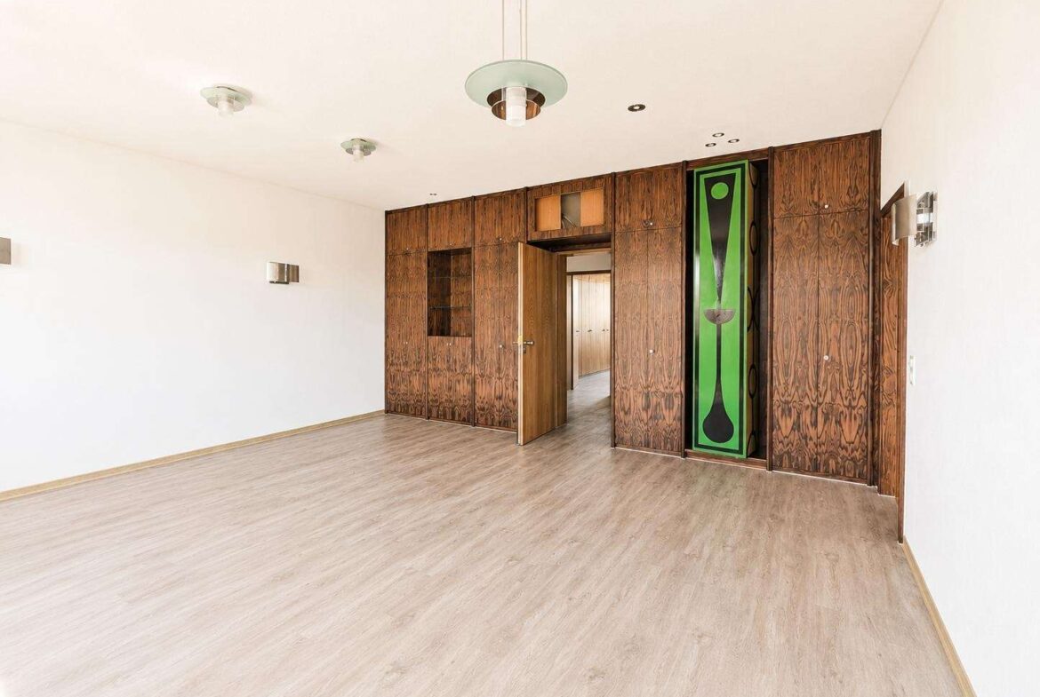 A spacious empty room with light wood flooring and dark wood built-in cabinets along the back wall, with an open doorway in the center-right area and a tall green decorative panel embedded in the cabinetry.