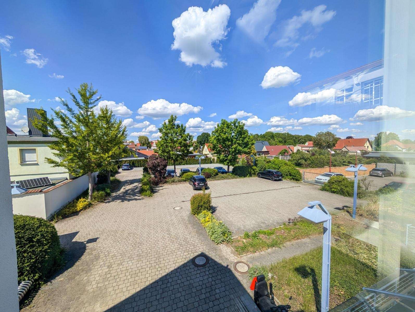 Parking lot with cobblestones, several cars, trees, and houses under a bright blue sky with fluffy white clouds (view from a window).