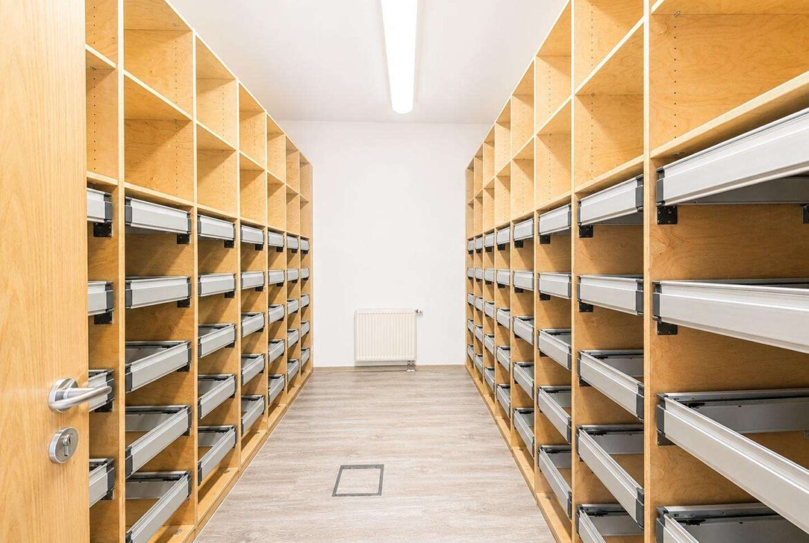 Empty storage room with tall wooden shelves and metal pull-out drawers along both walls, a central aisle.