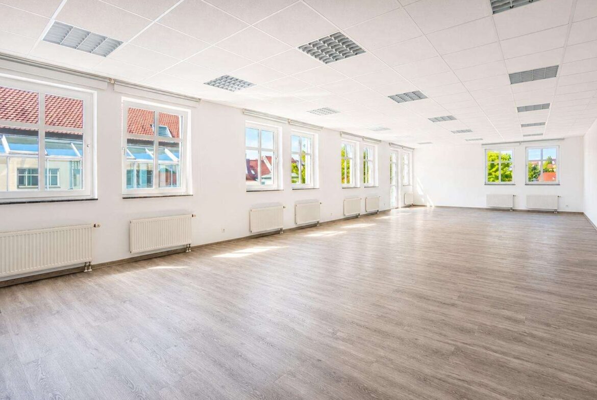 Bright, empty office or studio space with large windows, white walls, wood-tone flooring, and row of radiators under the windows