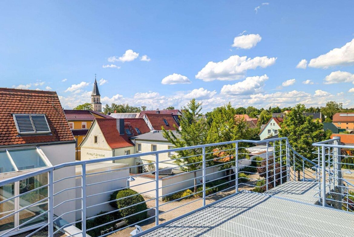 Rooftop balcony with metal railing overlooking a sunny town and a church steeple in the distance under a blue sky with fluffy clouds