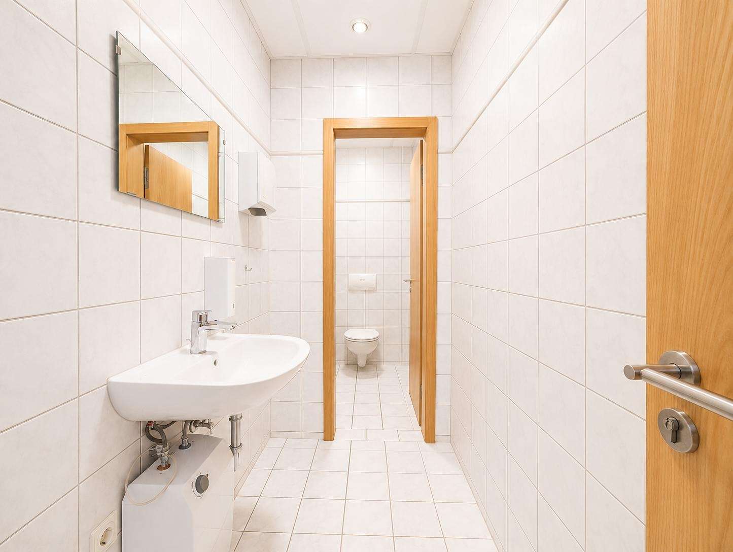 White-tiled bathroom with a wall-mounted sink, mirror, and a wooden doorway leading to a separate toilet at the back.
