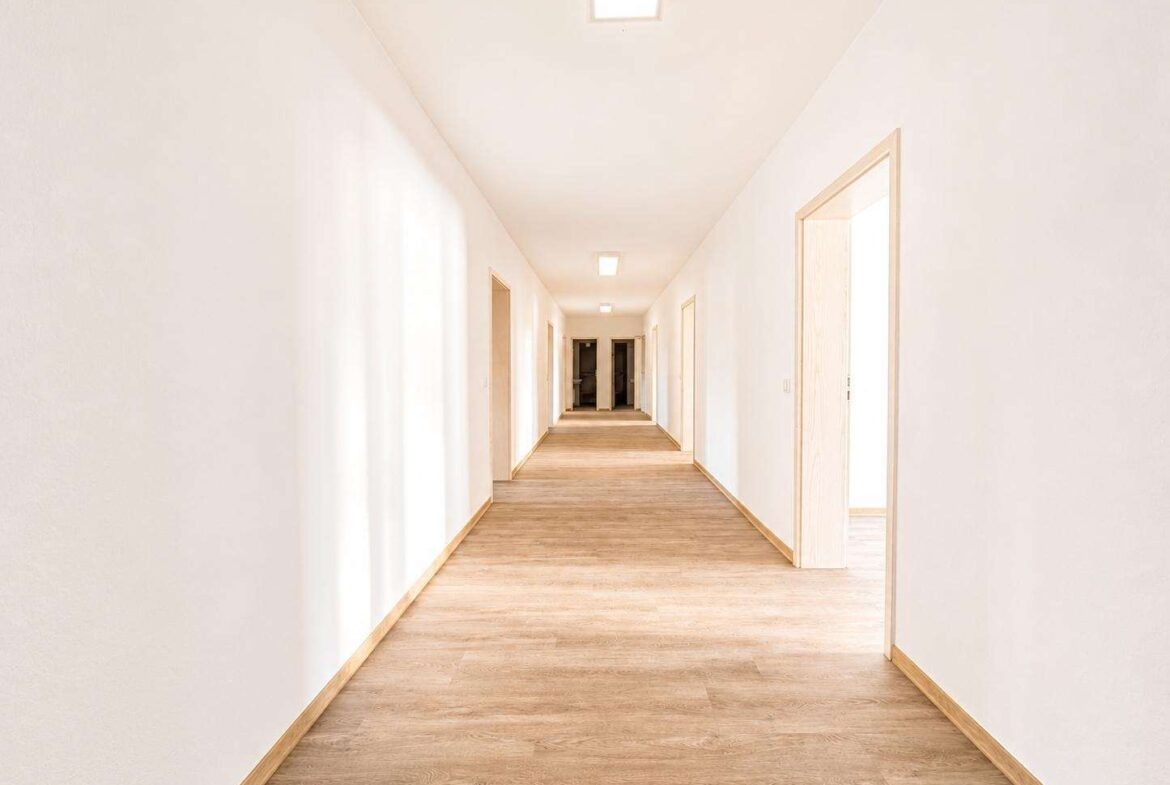 Bright empty hallway with white walls, wooden floor, and multiple doorways along both sides.