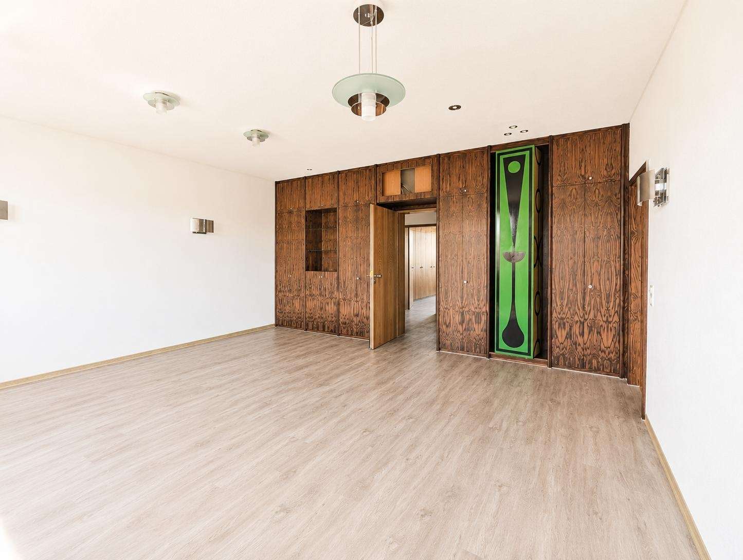 Empty living room with dark wood built-in cabinets along one wall, light wood flooring, and a green decorative panel near the cabinetry; bare walls and ceiling lights visible.