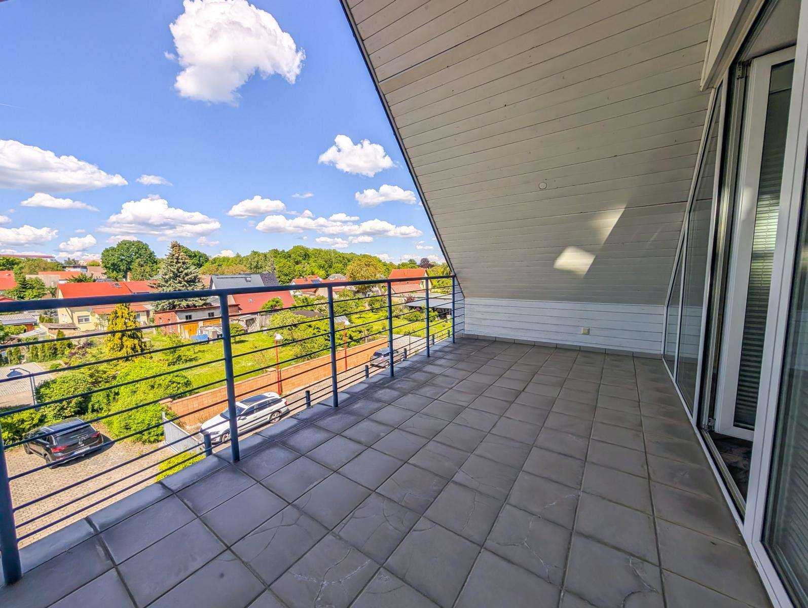 Balcony with gray tiled floor and blue railing, overlooking a sunny suburban neighborhood with trees and houses under a blue sky.
