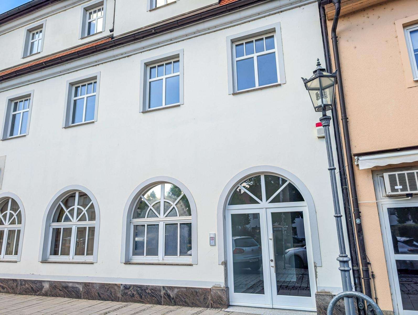 White building facade with large arched ground-floor windows and a glass door, street lamp on the right.