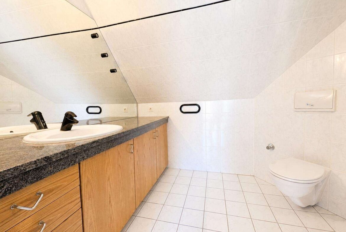 Bathroom with a double sink vanity, granite countertop, and light wood cabinets under a sloped, tiled ceiling. A wall-mounted toilet with a flush plate is visible to the right.