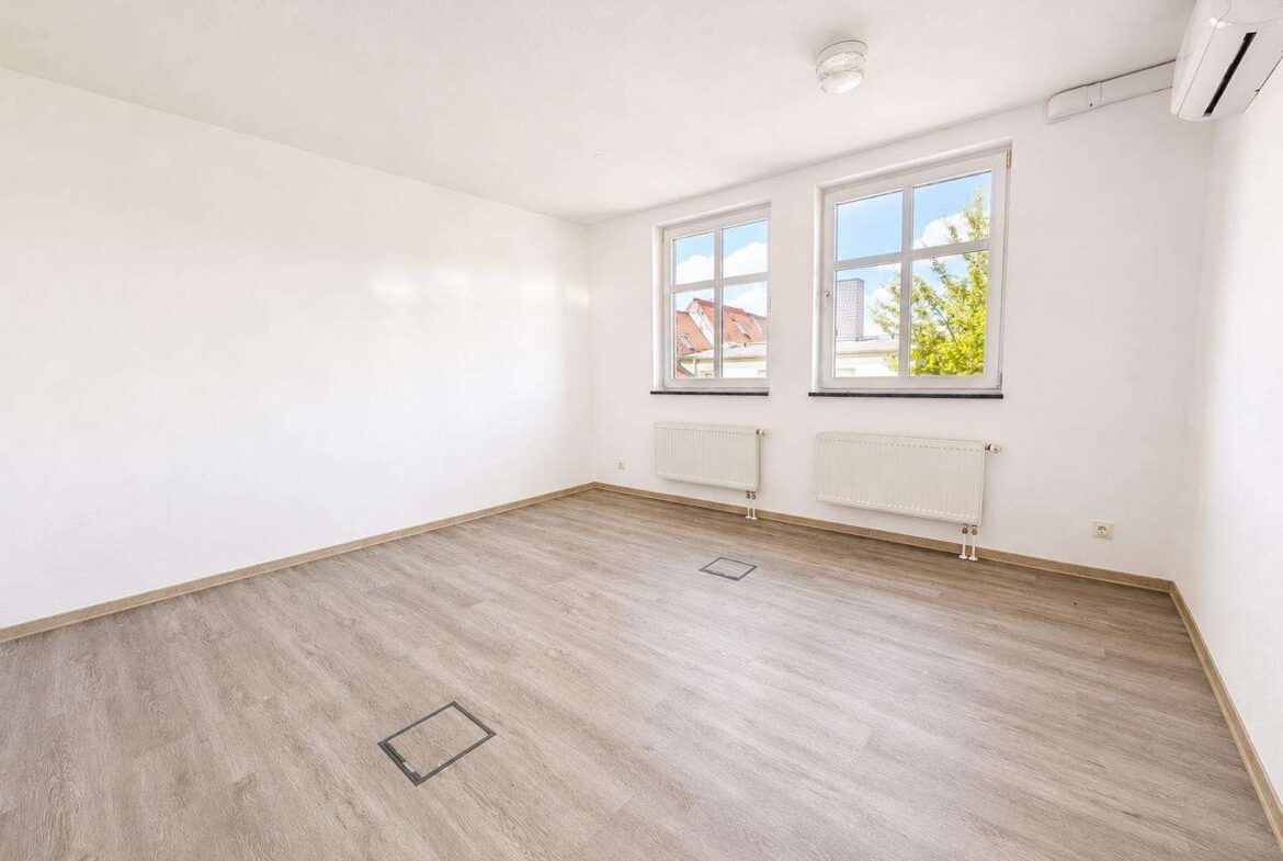 Bright empty room with light wood flooring, white walls, two windows, radiators beneath, and a wall-mounted air conditioner in the corner.