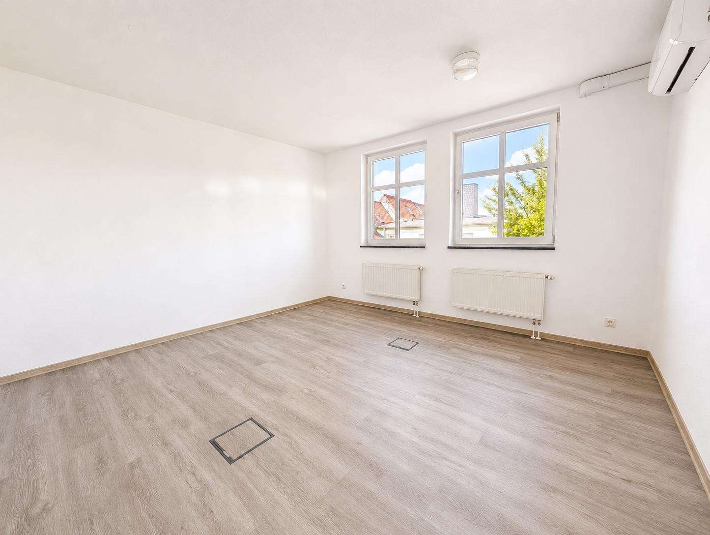 Bright empty room with light wood flooring, white walls, two windows, radiators beneath, and a wall-mounted air conditioner in the corner.