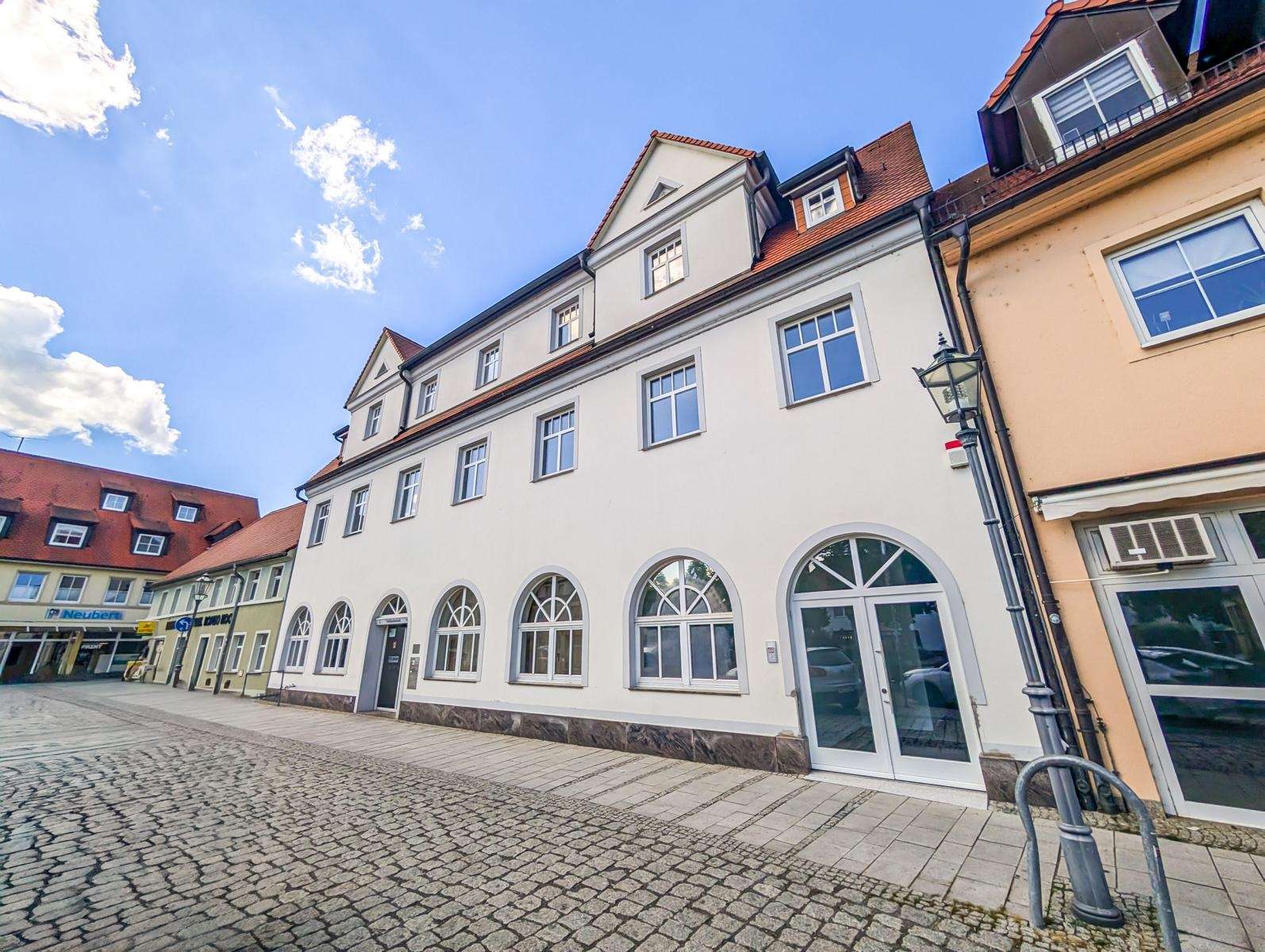 White multi-story building with arched ground-floor windows on a cobblestone street under a blue sky.
