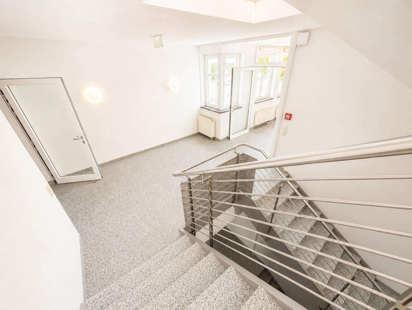 Bright stairwell in a modern building with metal handrails, gray speckled flooring, and a glass door to a sunlit hallway.