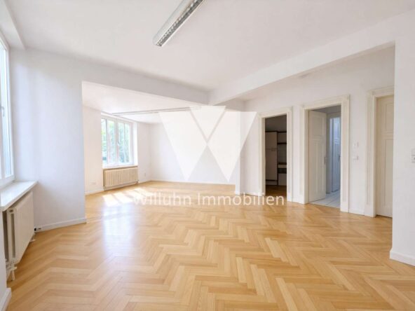 Bright, empty living room with herringbone wood floor, white walls, and large windows letting in natural light, plus radiators along the wall.