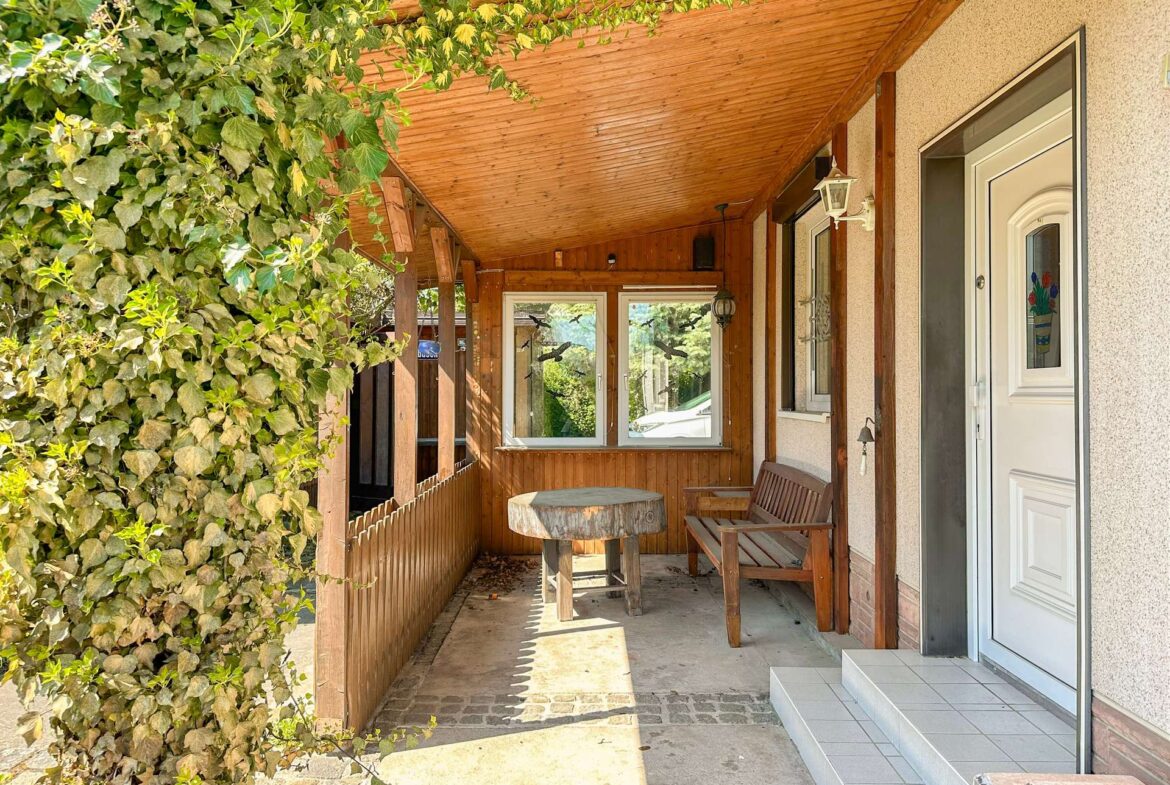 Covered porch with ivy-covered wall, wooden ceiling, and round tree-stump table with bench seating.