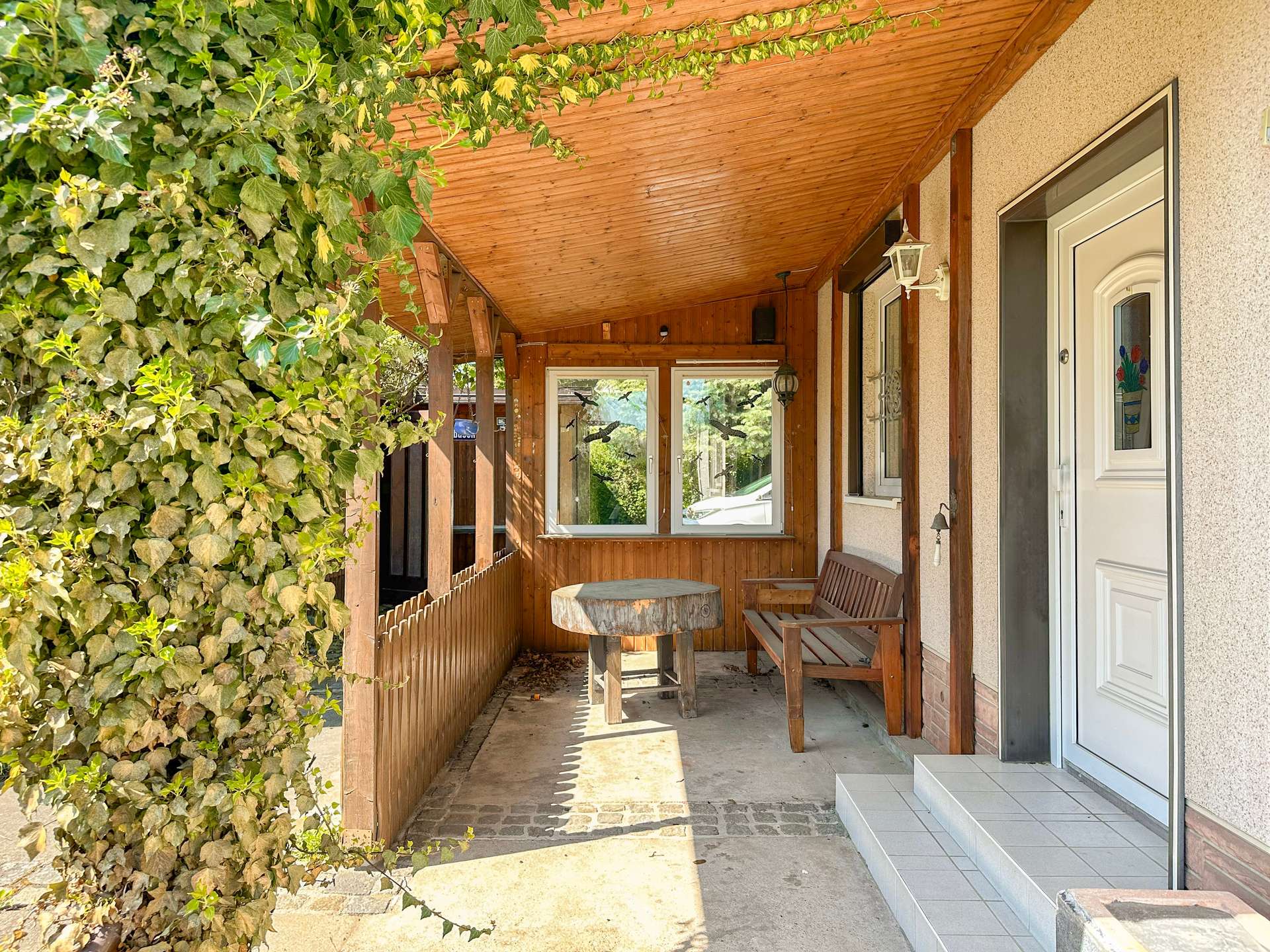 Covered porch with ivy-covered wall, wooden ceiling, and round tree-stump table with bench seating.