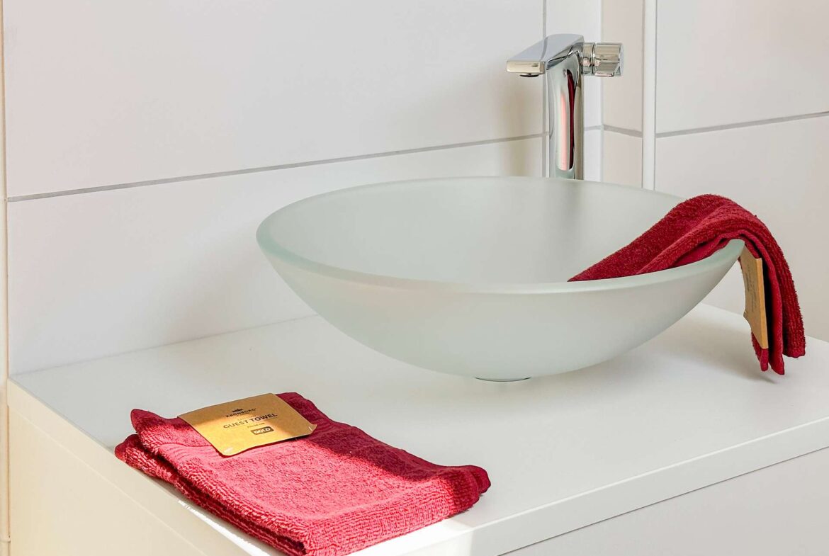 White vessel sink with a tall chrome faucet on a white vanity; a red towel rests on the counter, with a guest towel tag nearby.