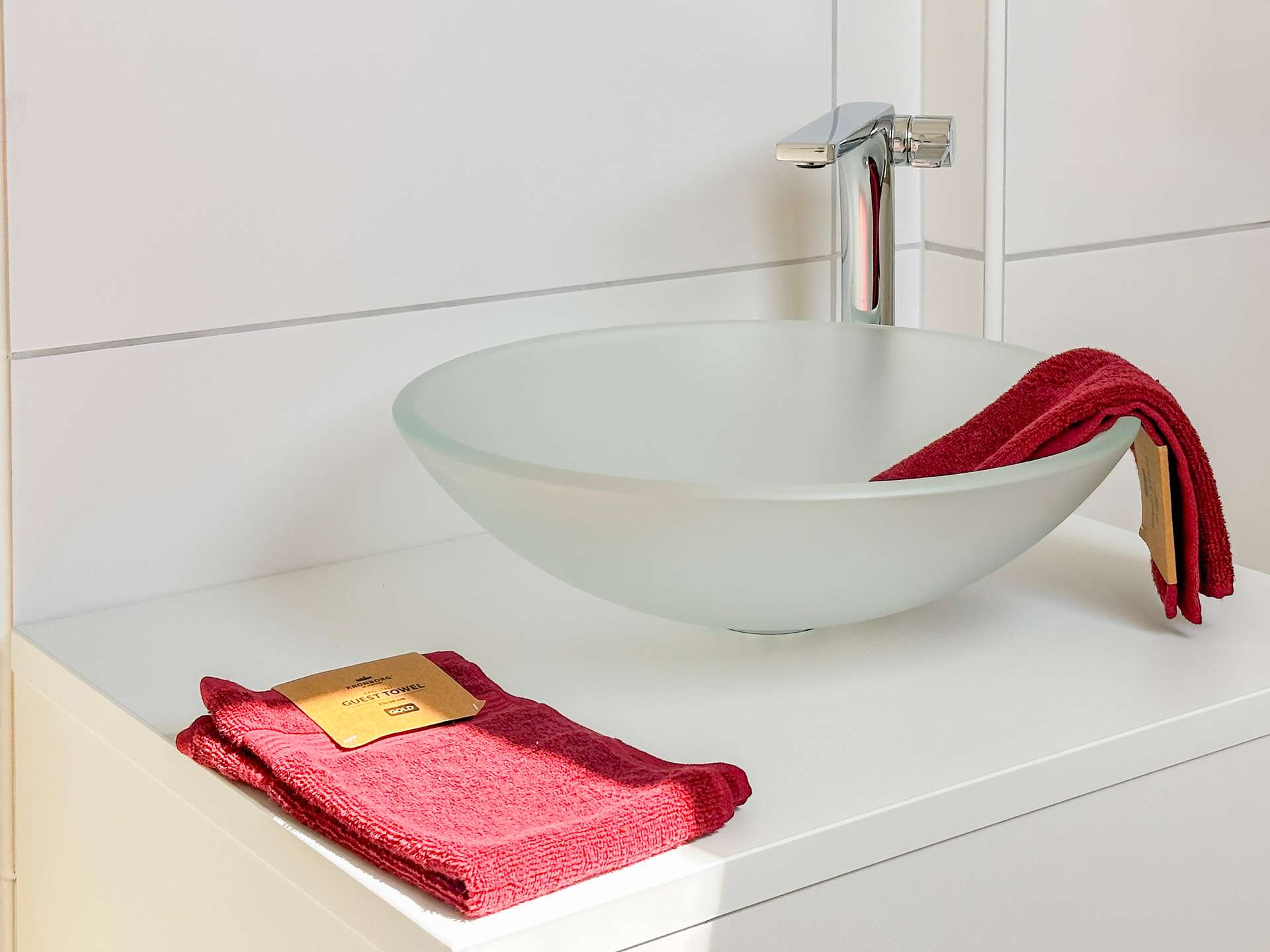 White vessel sink with a tall chrome faucet on a white vanity; a red towel rests on the counter, with a guest towel tag nearby.