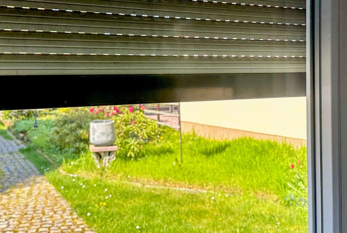 Partially closed metal roller shutter above a window; view of a garden with a stone path, birdbath, and green plants outside.